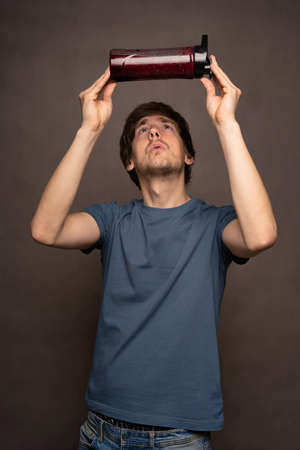 Young handsome tall slim white man with brown hair observing and holding smoothie bottle over his head in grey shirt on grey backgroundの写真素材