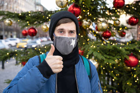 Young handsome white man with brown hair in blue jacket with black winter hat making thumb up with...