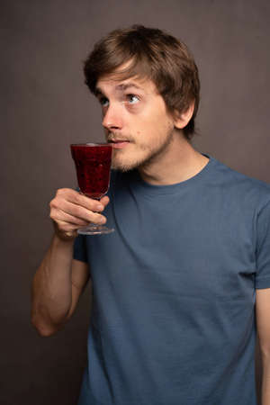 Young handsome tall slim white man with brown hair smelling wine cup curiously in grey shirt on grey backgroundの写真素材