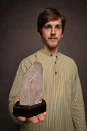 Young handsome tall slim white man with brown hair presenting big crystal in striped shirt on grey backgroundの写真素材