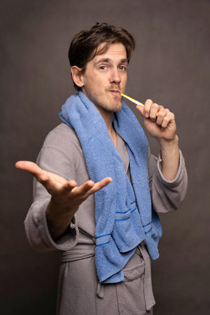 Young handsome tall slim white man with brown hair brushing his teeth offering his right hand in grey bathrobe on grey backgroundの写真素材