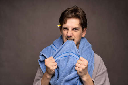 Young handsome tall slim white man with brown hair angrily biting into blue towel in grey bathrobe on grey backgroundの写真素材