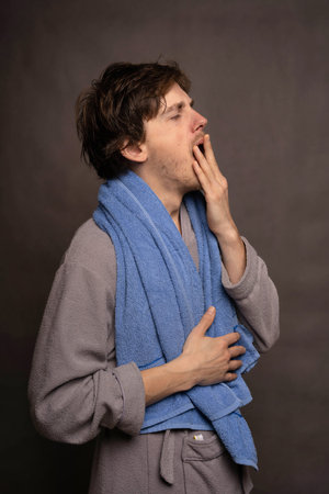 Young handsome tall slim white man with brown hair yawning with towel on shoulders in grey bathrobe on grey backgroundの写真素材