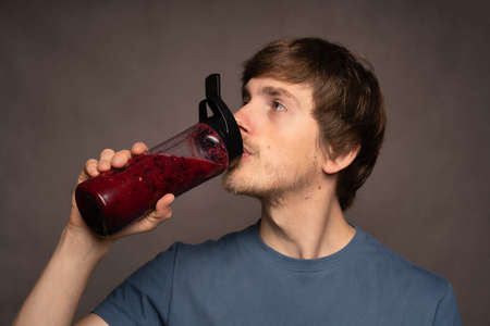 Young handsome tall slim white man with brown hair drinking from smoothie bottle in grey shirt on grey backgroundの写真素材