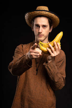 Young handsome tall slim white man with brown hair holding bananas pointing at you with brown shirt and straw hat on black backgroundの写真素材