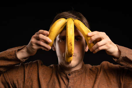 Young handsome tall slim white man with brown hair with bananas on head with brown shirt on black backgroundの写真素材