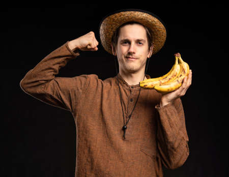Young handsome tall slim white man with brown hair flexing his muscles holding bananas with brown shirt and straw hat on black backgroundの写真素材