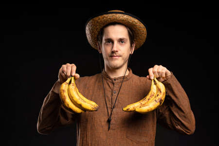 Young handsome tall slim white man with brown hair holding bananas looking into camera with brown shirt and straw hat on black backgroundの写真素材