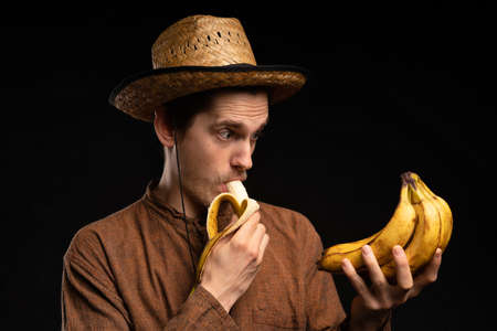 Young handsome tall slim white man with brown hair biting banana holding bunch of bananas with brown...