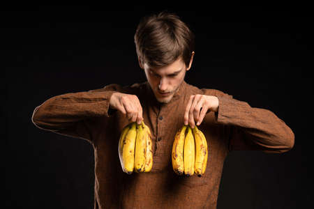 Young handsome tall slim white man with brown hair holding bananas on chest with brown shirt on black backgroundの写真素材
