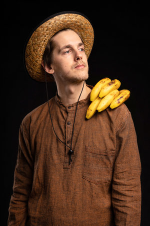 Young handsome tall slim white man with brown hair bananas on shoulder with brown shirt and straw hat on black backgroundの写真素材