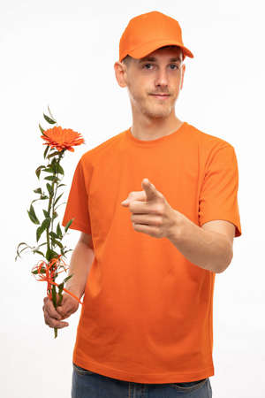 Young handsome tall slim white man with brown hair holding orange flower pointing at you with orange shirt isolated on white backgroundの写真素材
