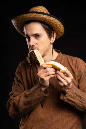 Young handsome tall slim white man with brown hair taking banana peel off with teeth with brown shirt and straw hat on black backgroundの写真素材