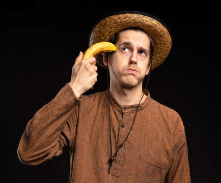 Young handsome tall slim white man with brown hair pointing banana to his head with brown shirt and straw hat on black backgroundの写真素材