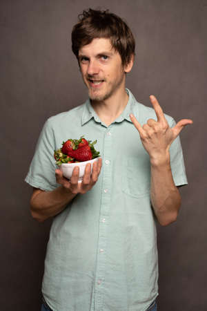 Young handsome tall slim white man with brown hair holding strawberries making horns sign in light blue shirt on grey backgroundの写真素材