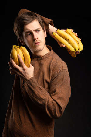 Young handsome tall slim white man with brown hair posing with bananas looking serious with brown shirt on black backgroundの写真素材
