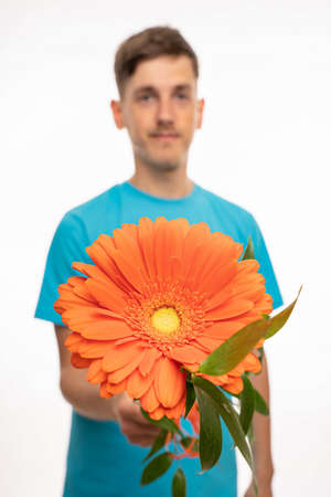 Young handsome tall slim white man with brown hair offering orange flower to you with blue shirt isolated on white backgroundの写真素材