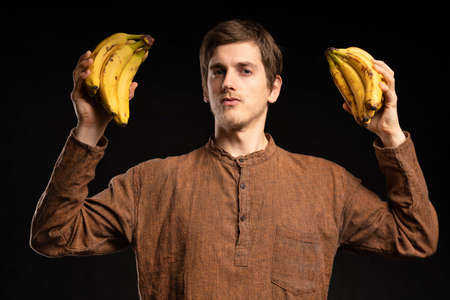 Young handsome tall slim white man with brown hair holding bananas looking strong with brown shirt on black backgroundの写真素材