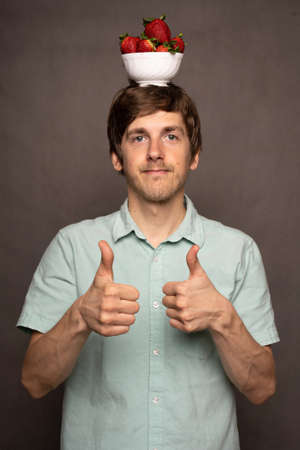 Young handsome tall slim white man with brown hair balancing strawberries on head giving thumbs up...