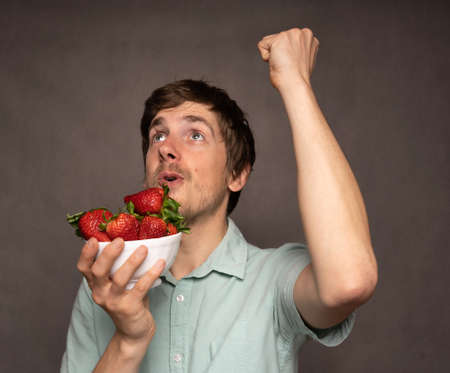 Young handsome tall slim white man with brown hair holding strawberries cheering in light blue shirt on grey backgroundの写真素材