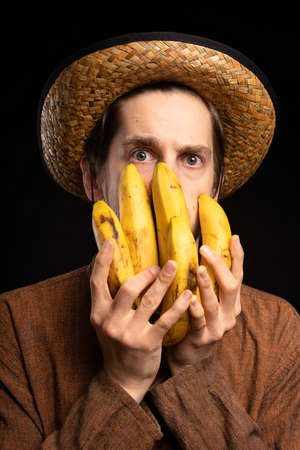 Young handsome tall slim white man with brown hair covering face with bananas with brown shirt and straw hat on black backgroundの写真素材