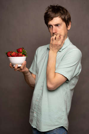 Young handsome tall slim white man with brown hair looking worried holding strawberries in light blue shirt on grey backgroundの写真素材