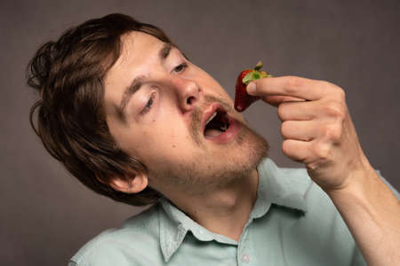 Young handsome tall slim white man with brown hair about to eat a strawberry in light blue shirt on grey backgroundの写真素材