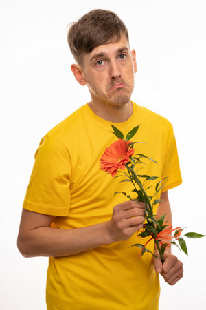 Young handsome tall slim white man with brown hair looking sad holding orange flower in yellow shirt isolated on white backgroundの写真素材