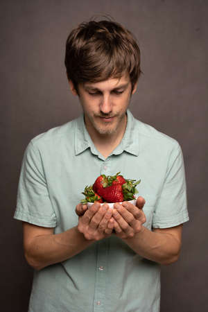 Young handsome tall slim white man with brown hair holding strawberries looking down in light blue shirt on grey backgroundの写真素材
