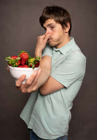 Young handsome tall slim white man with brown hair holding strawberries and his nose arms crossed in light blue shirt on grey backgroundの写真素材
