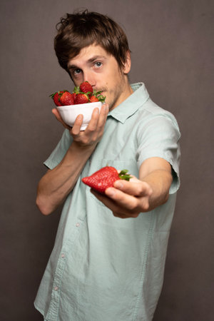 Young handsome tall slim white man with brown hair sniffing strawberries holding one in light blue shirt on grey backgroundの写真素材