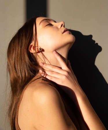 Portrait in sunset light of woman with dark hair and white skin isolated on beige with closed eyes and hand on her neckの写真素材