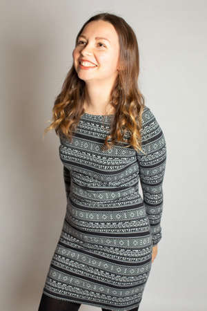 Smiling young woman in warm striped dress isolated on light grey holding hands on her back