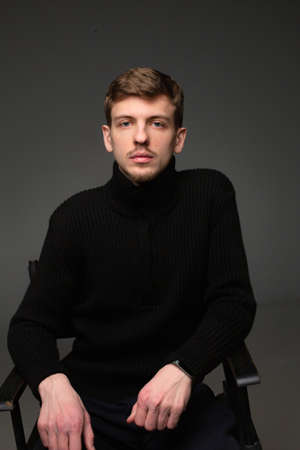 Young blond european man with short beard and mustache sitting isolated on dark grey in total black outfitの写真素材