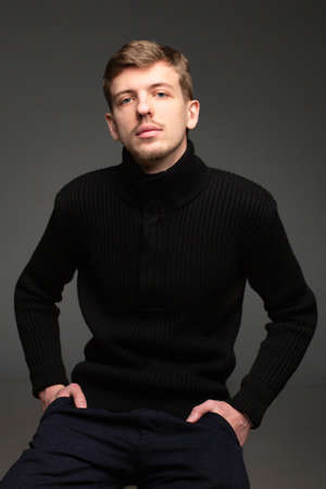 Young blond european man with short beard and mustache isolated on dark grey in total black with hands in pocketsの写真素材