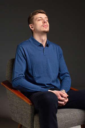 Attractive young blond european man with short beard and mustache isolated on black sitting in armchairの写真素材