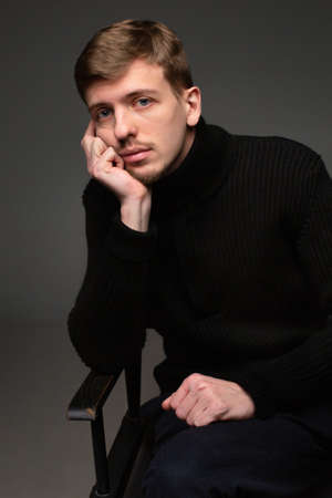 Young blond european man with short beard and mustache sitting isolated on dark grey holding hand near his faceの写真素材