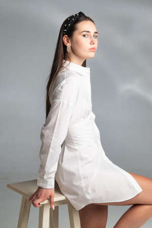 Young woman with dark hair in white long sleeve blouse isolated on grey sitting on chairの写真素材