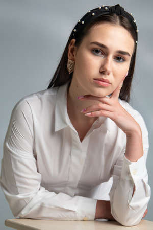 Young woman with dark hair in white long sleeve blouse leaning on chair isolated on grey holding hand near her faceの写真素材