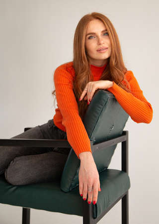 Woman with long red hair in orange jumper isolated on beige sitting on chairの写真素材