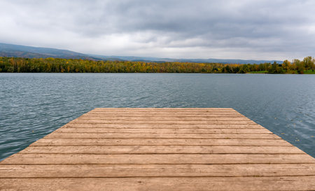 Wooden pier extending into a calm lake surrounded by autumn trees with colorful foliage. Peaceful natural landscape under cloudy sky, ideal for relaxation, travel, or seasonal background.の写真素材
