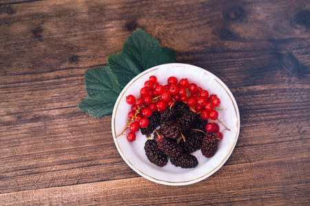 red currant berries and mulberries on a white plate on wooden background