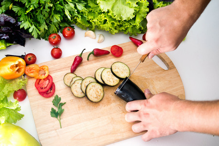 A man prepares a dinner of vegetables, cuts eggplants, tomatoes, peppers, lettuce, greens. Useful vegetarian foodの写真素材