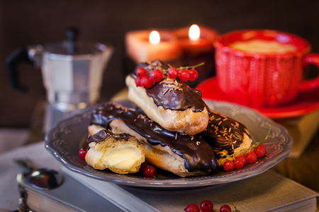 Chocolate eclairs with custard cream and cup of coffee on cozy candles evening background