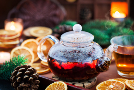 Glass teapot of hot black tea on cozy background with dried oranges and candlesの写真素材