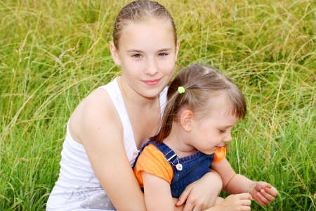 Sisters on a farm  Two children cute young girl teenager and cute little girl are sitting on green meadow and smile enjoying the summer dayの写真素材