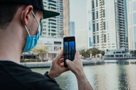 Person tourist with medical mask outdoor holding phone and taking pictures and videos of the city on the modern smartphone cameraの写真素材