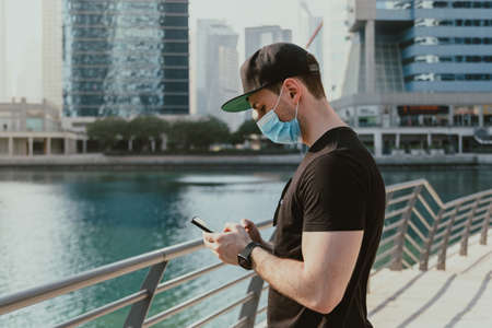 Person tourist with medical mask holding and using  phone by typing messages while standing outdoor on the street with urban background and skyscrapersの写真素材