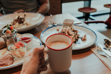 Person holding a cappuccino cup in front of breakfast table with assorted food itemsの写真素材