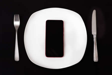 Food delivery concept. Top view on smartphone laying on plate on black table. Smart phone black screen on white plate, fork and knife, black background table. Take away restaurant meal onlineの写真素材